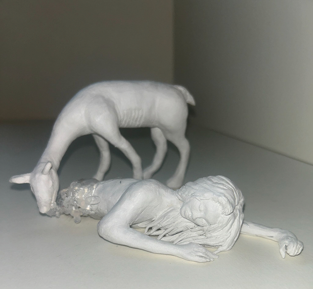 Sculpture of the upper half of a dead woman laying on the ground with a deer drinking from the fluids draining out of the torso.